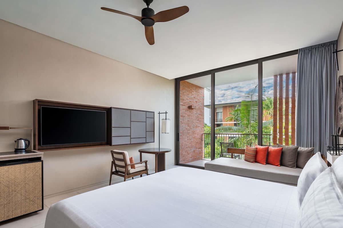 Une chambre d’hôtel avec un lit, un téléviseur fixé au mur, un ventilateur de plafond, une petite table avec une chaise, et une vue sur l’extérieur.