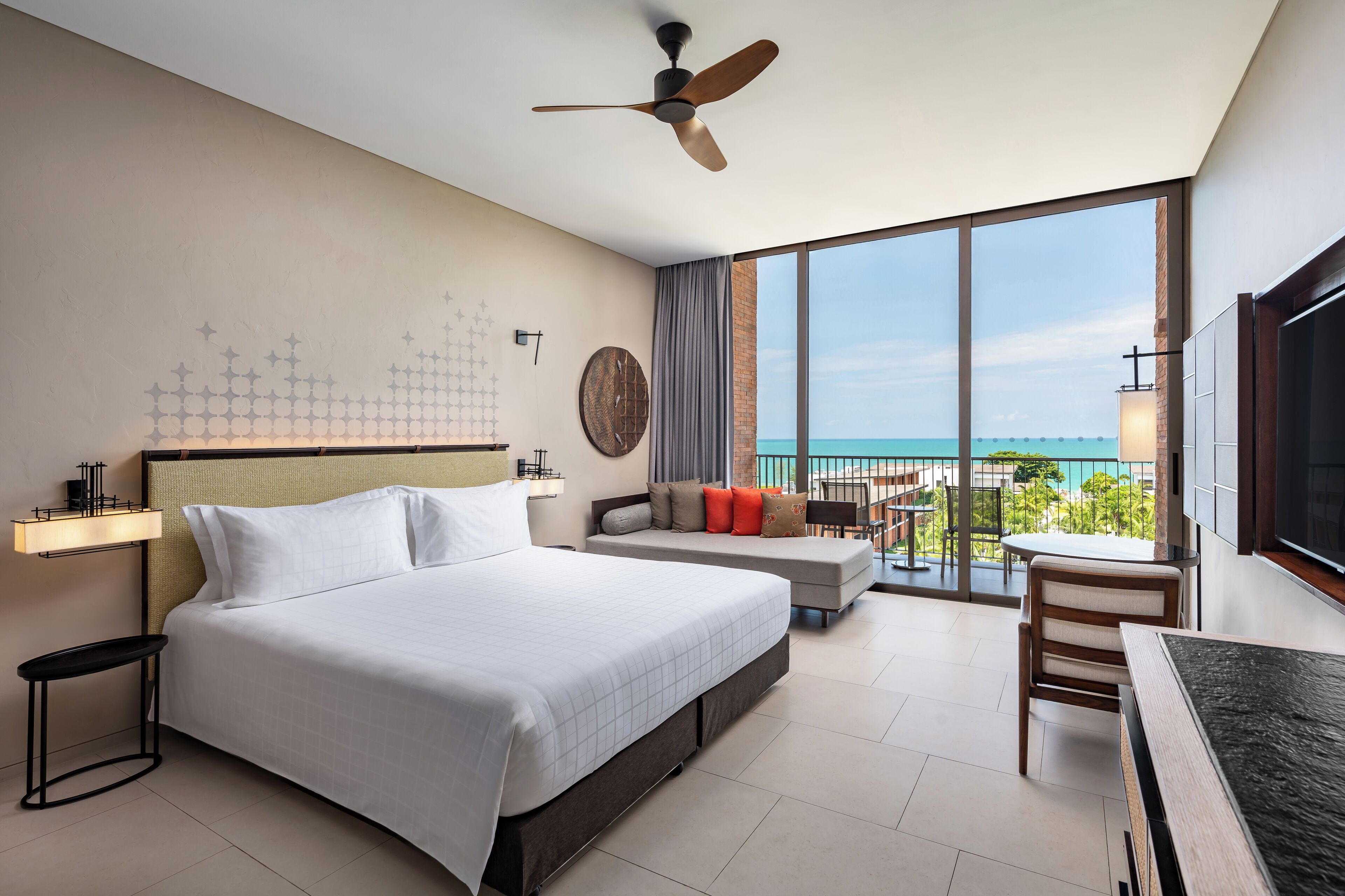 A hotel room with a large bed, a ceiling fan, a balcony with a view of the ocean, and a dining area.