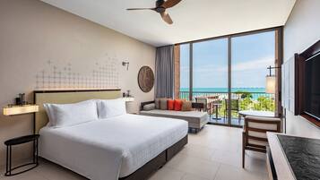A hotel room with a large bed, a ceiling fan, a balcony with a view of the ocean, and a dining area.
