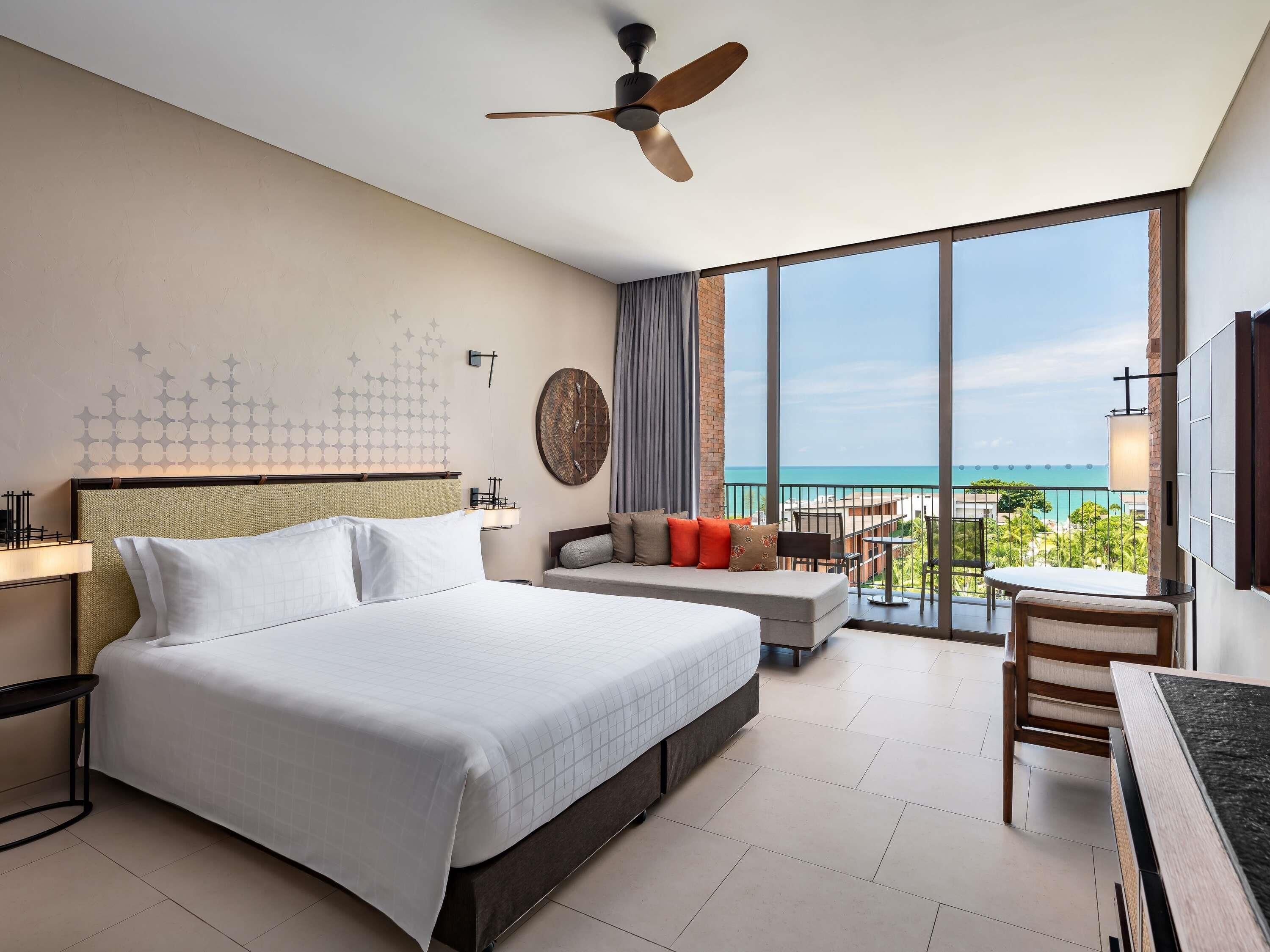 Habitación de hotel con cama, escritorio, silla, balcón con vista al océano y ventilador de techo.