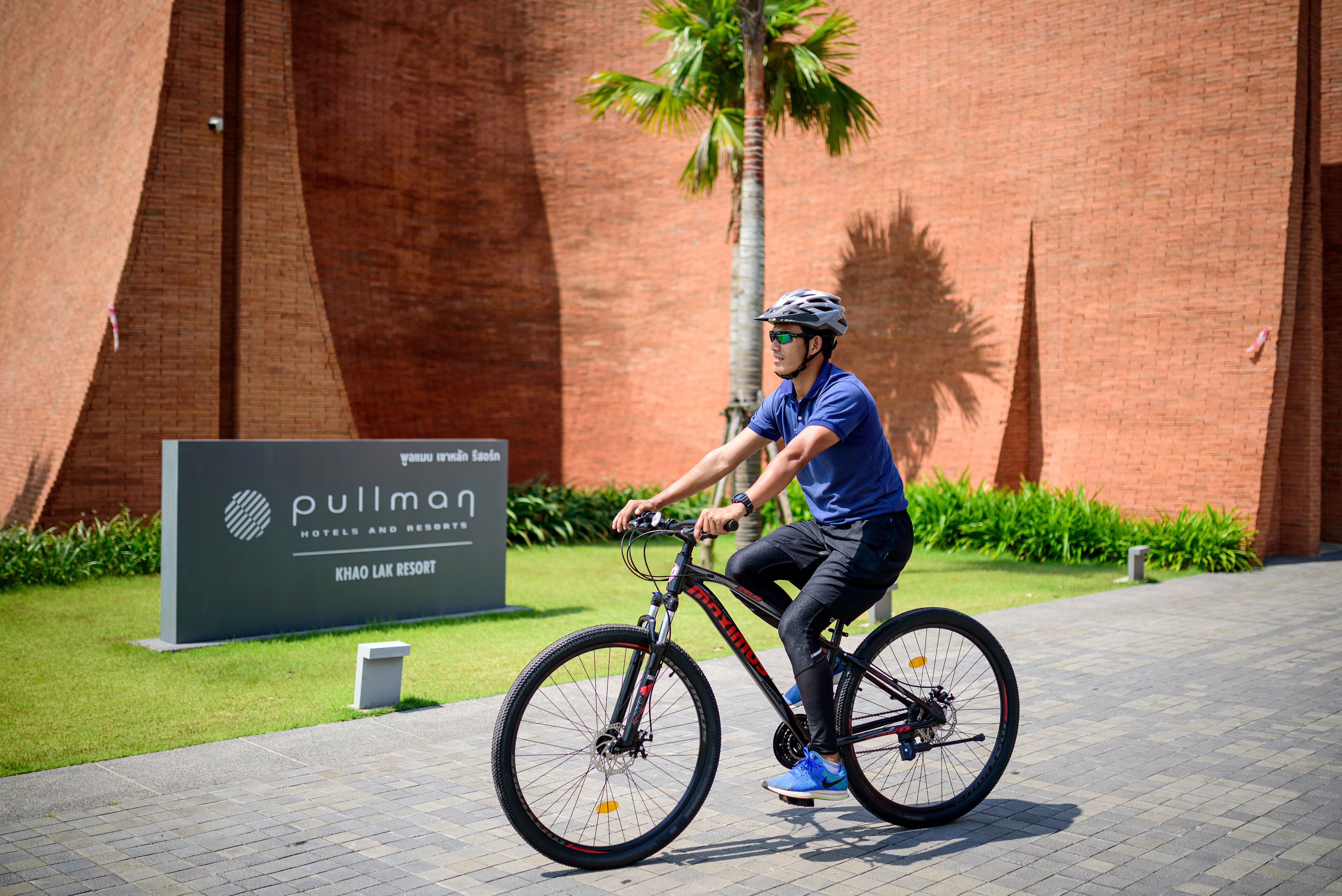 A cyclist riding a bike in front of a building with a sign that reads "Pullman Hotels and Resorts Khao Lak Resort".
