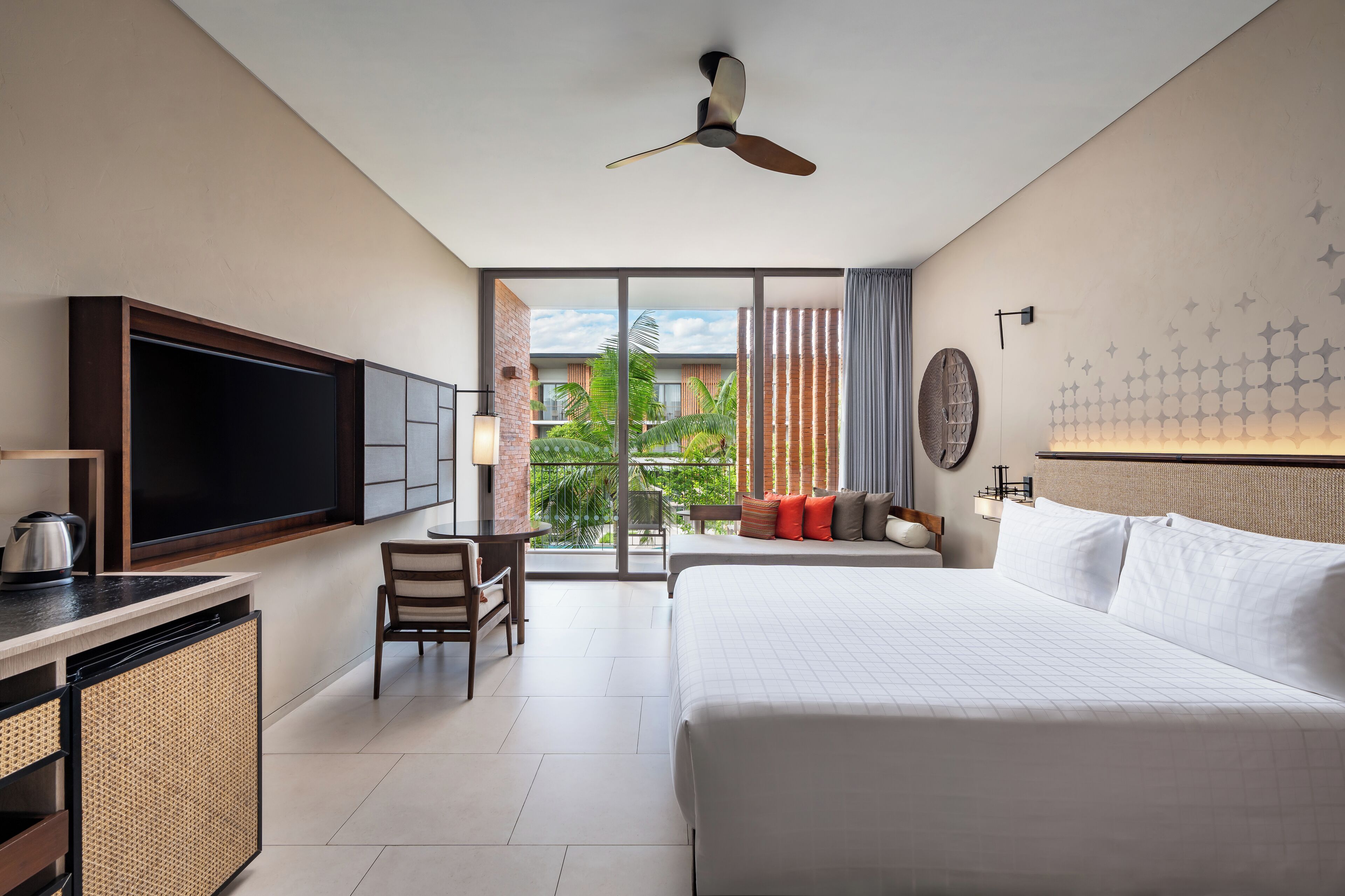 Une chambre d’hôtel avec un grand lit, un bureau avec une télévision, un balcon donnant sur un espace verdoyant et un ventilateur de plafond.