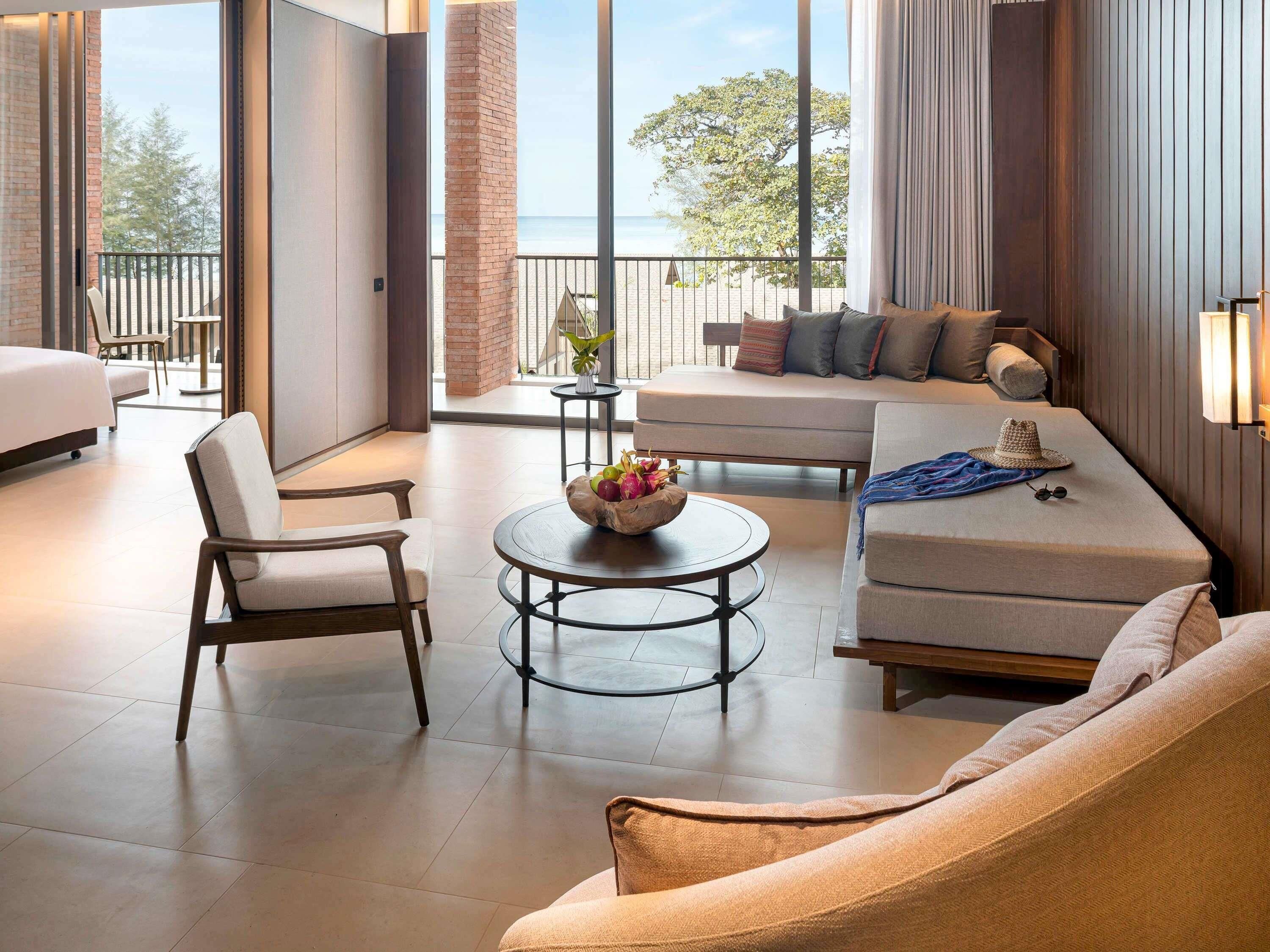 A modern hotel room with a balcony, a sofa, a chair, a round table with fruit, and a view of the beach.