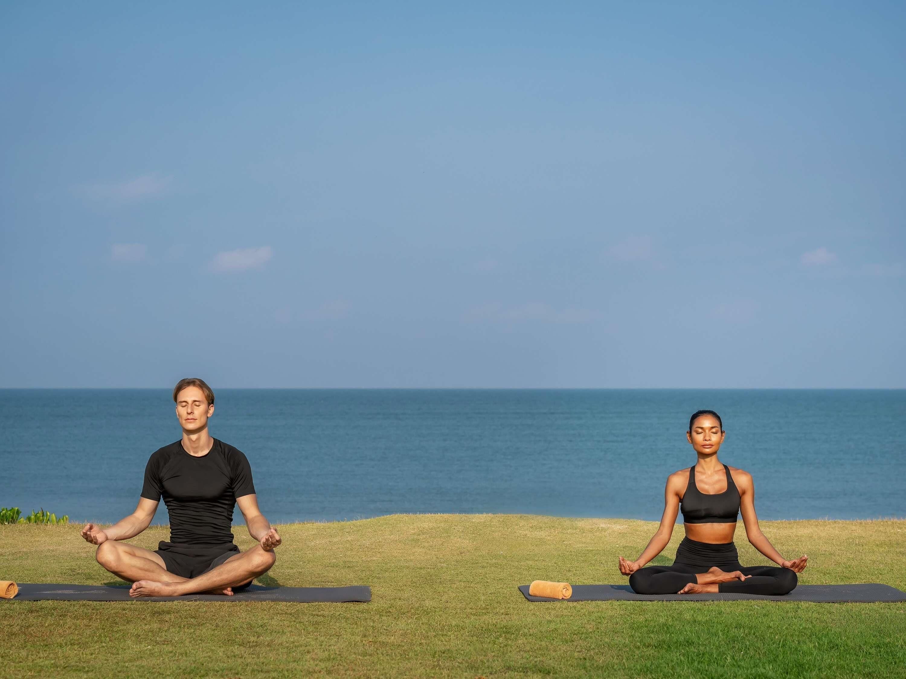 Two individuals meditating on yoga mats by a body of water.