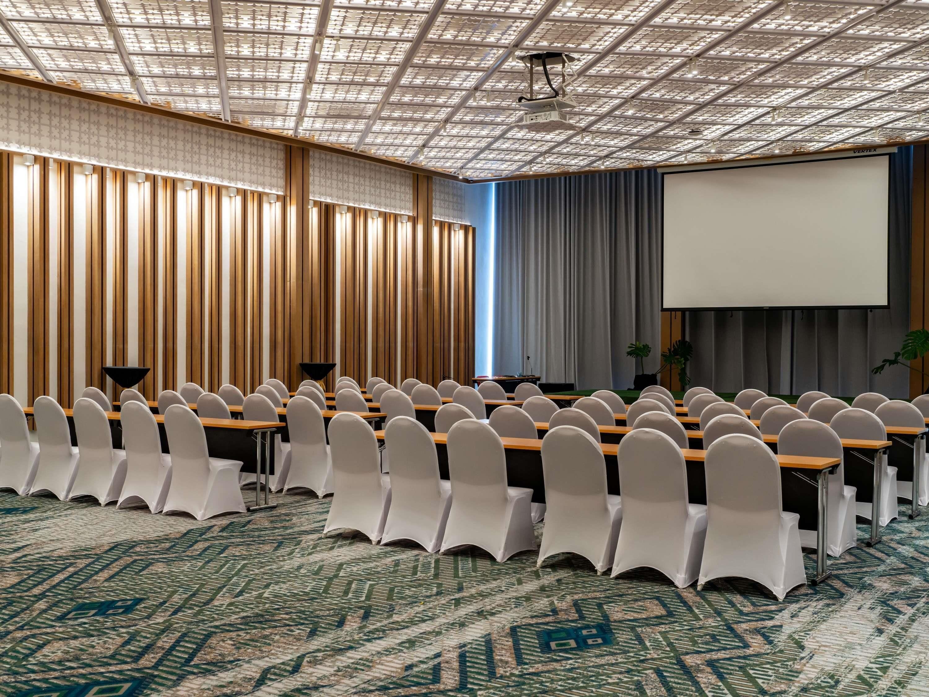Un salón de conferencias con filas de sillas blancas, una pantalla de proyector y una alfombra estampada.