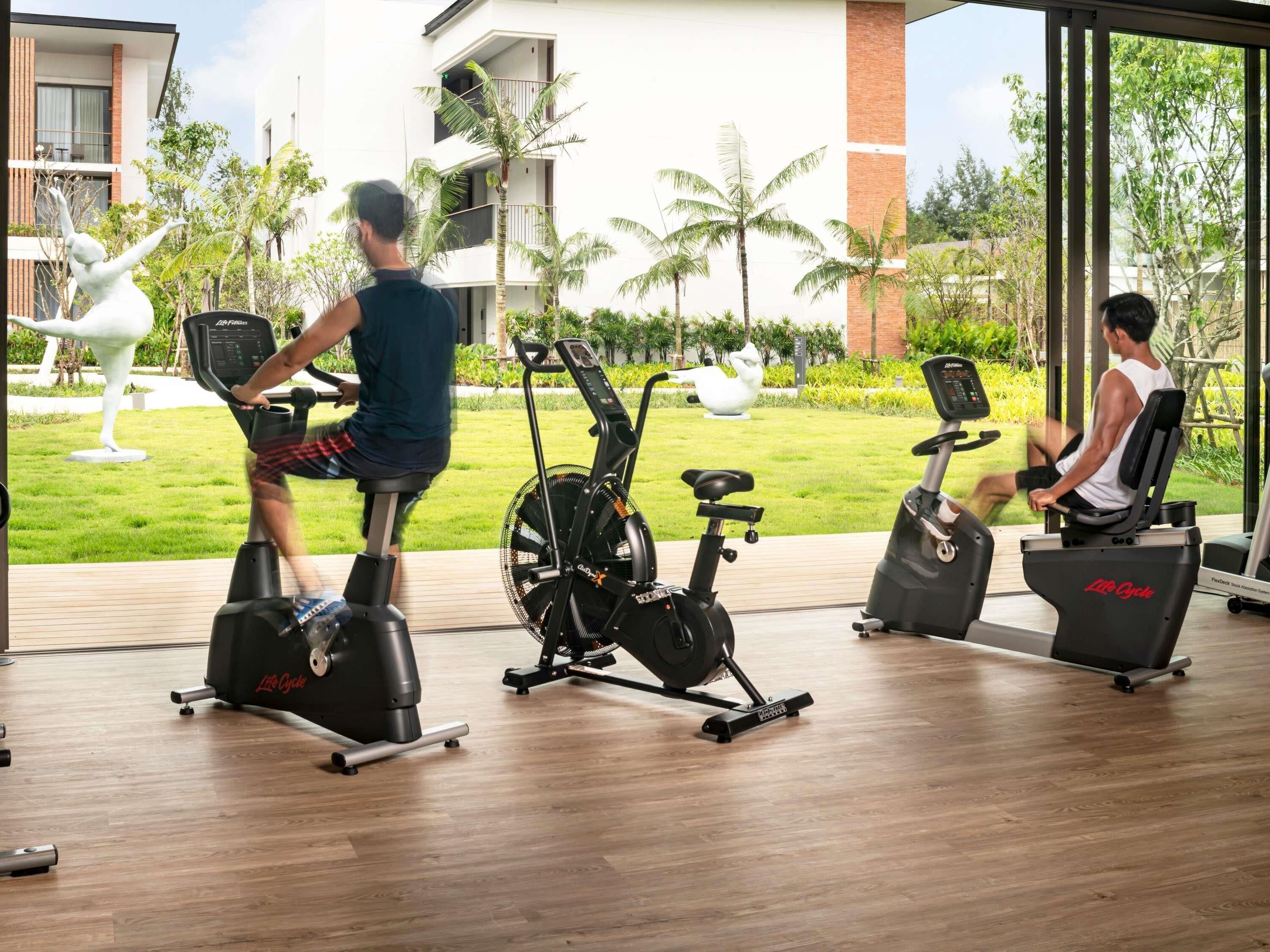 Ginásio com bicicletas ergométricas e equipamentos elípticos, grandes janelas e vista para um edifício e áreas verdes.