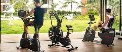 Ginásio com bicicletas ergométricas e equipamentos elípticos, grandes janelas e vista para um edifício e áreas verdes.