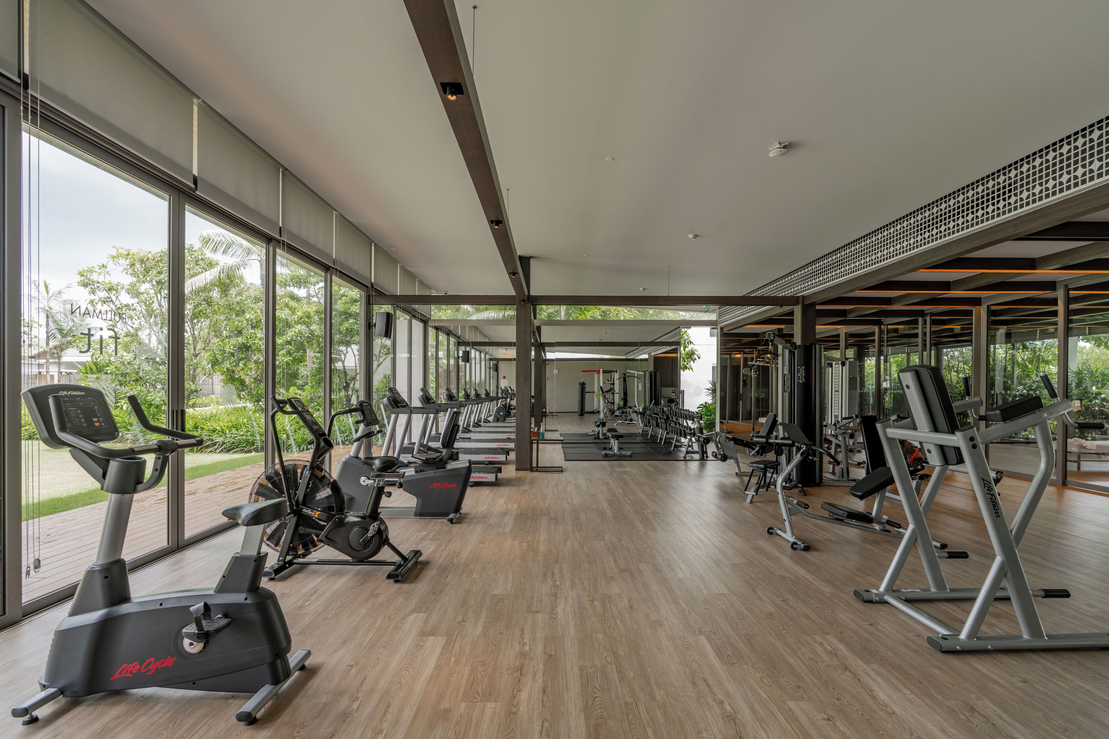 A modern gym with cardio machines, weight stations, and large windows offering views of greenery.