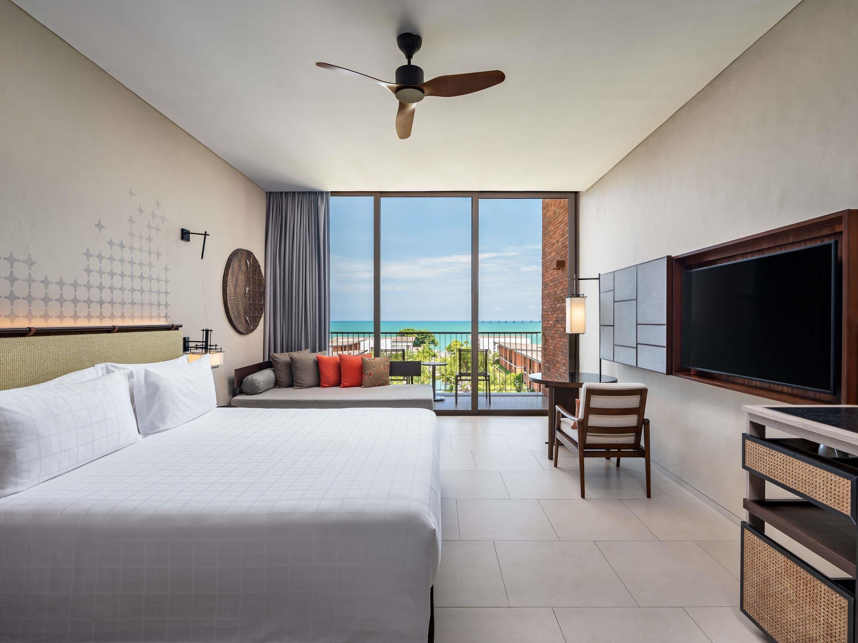 Een hotelkamer met een bed, bureau, stoel, televisie en balkon met uitzicht op de oceaan.