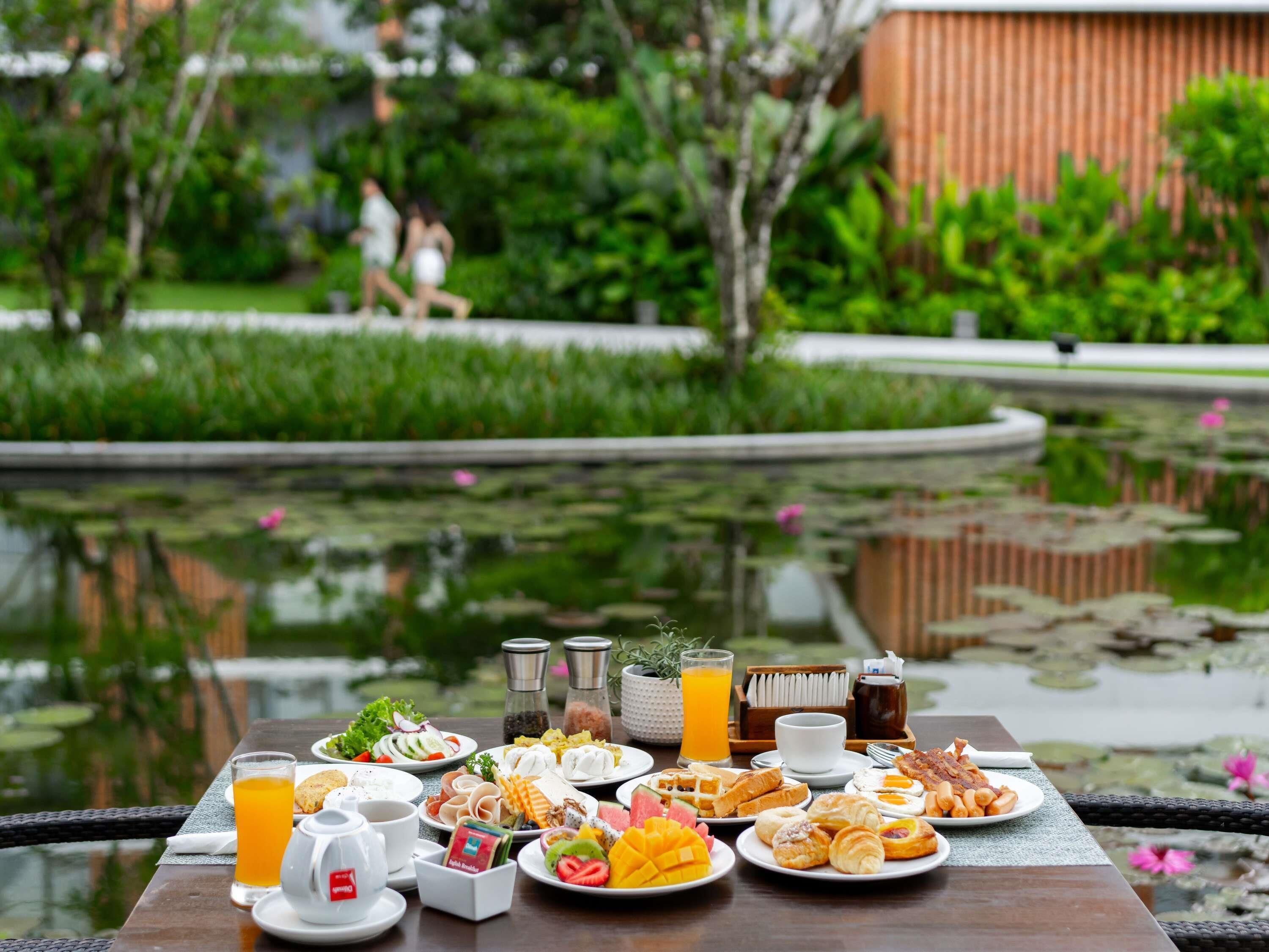 A breakfast buffet with various dishes and beverages on a table by a pond.