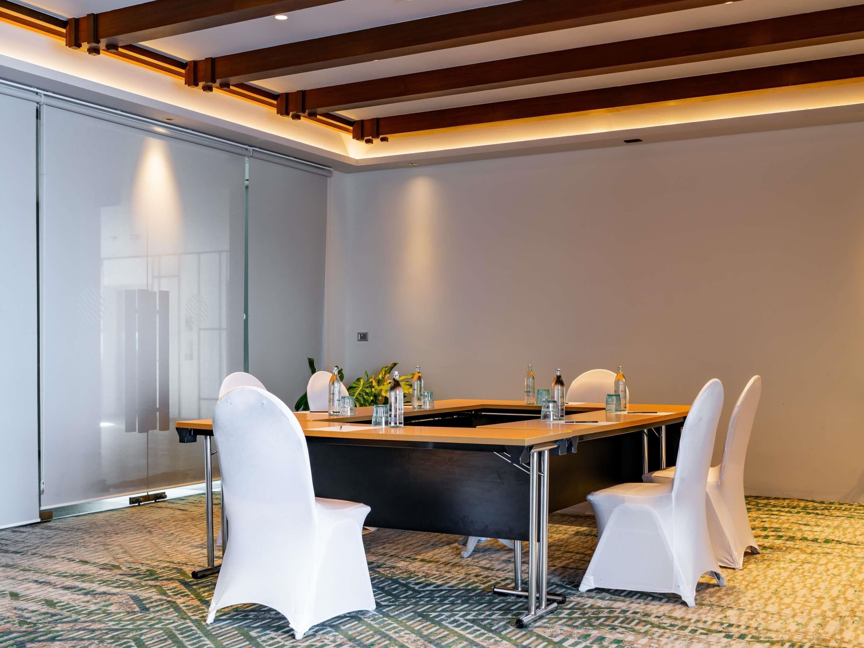 Un salón de conferencias con una larga mesa, sillas blancas y un panel de vidrio.