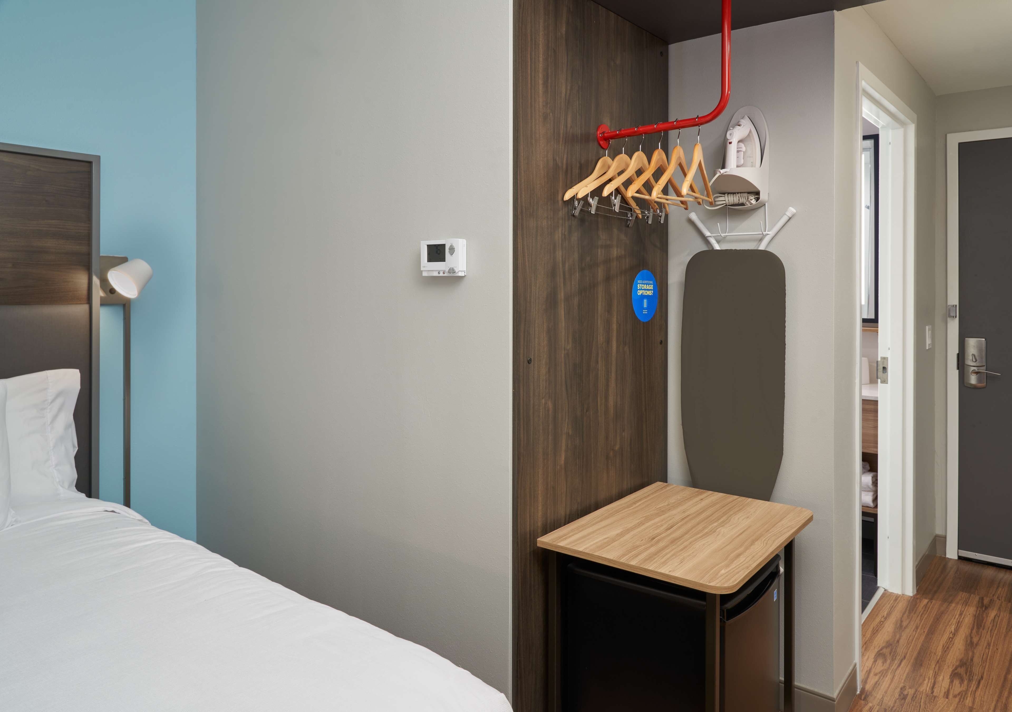 Quarto de hotel com uma cama, uma mesinha, aquecedor fixado na parede, cabideiro e uma porta que dá para um banheiro.