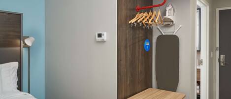 Habitación de hotel con cama, una mesita, un calentador en la pared, perchero y puerta que da al baño.
