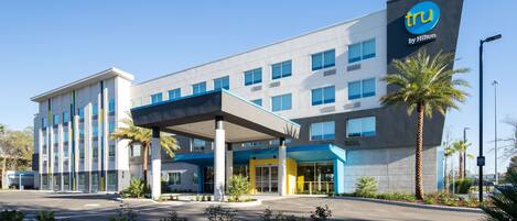 A modern hotel with a prominent 'tru by Hilton' sign, a covered entrance, and a landscaped front yard with palm trees.