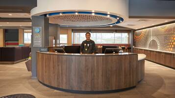 A modern hotel reception area with a curved wooden reception desk, a digital display, and a large window offering a view of the outdoors.