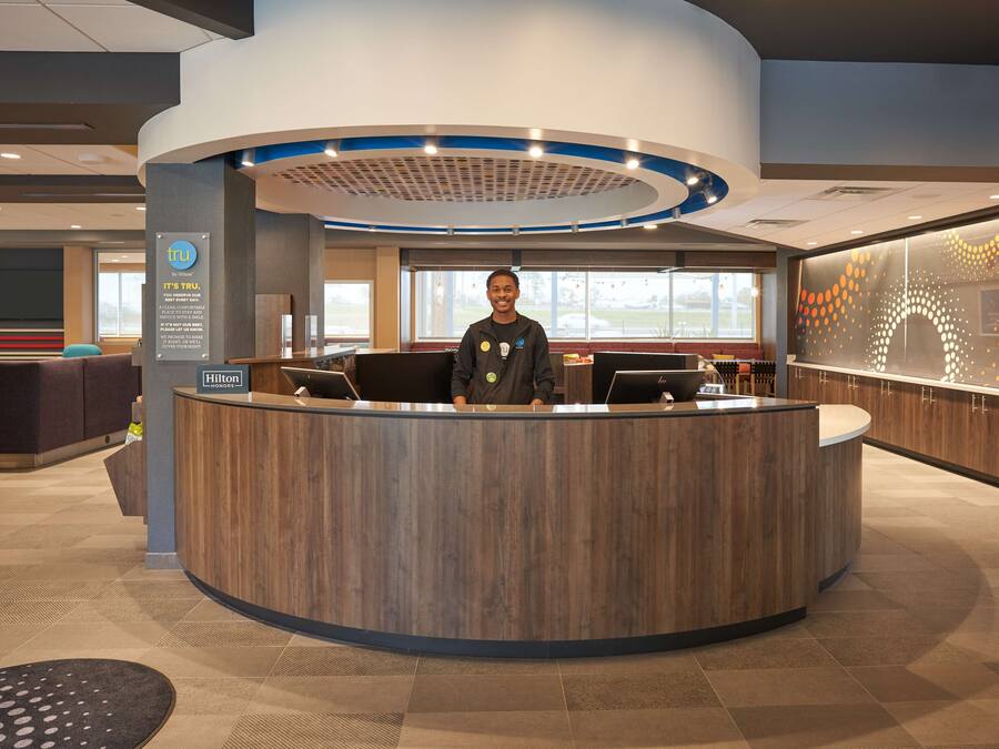 A modern hotel reception area with a curved wooden reception desk, a digital display, and a large window offering a view of the outdoors.