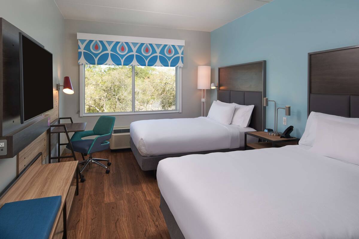 A hotel room with two beds, a desk, a chair, a TV, and a window with a view of trees.