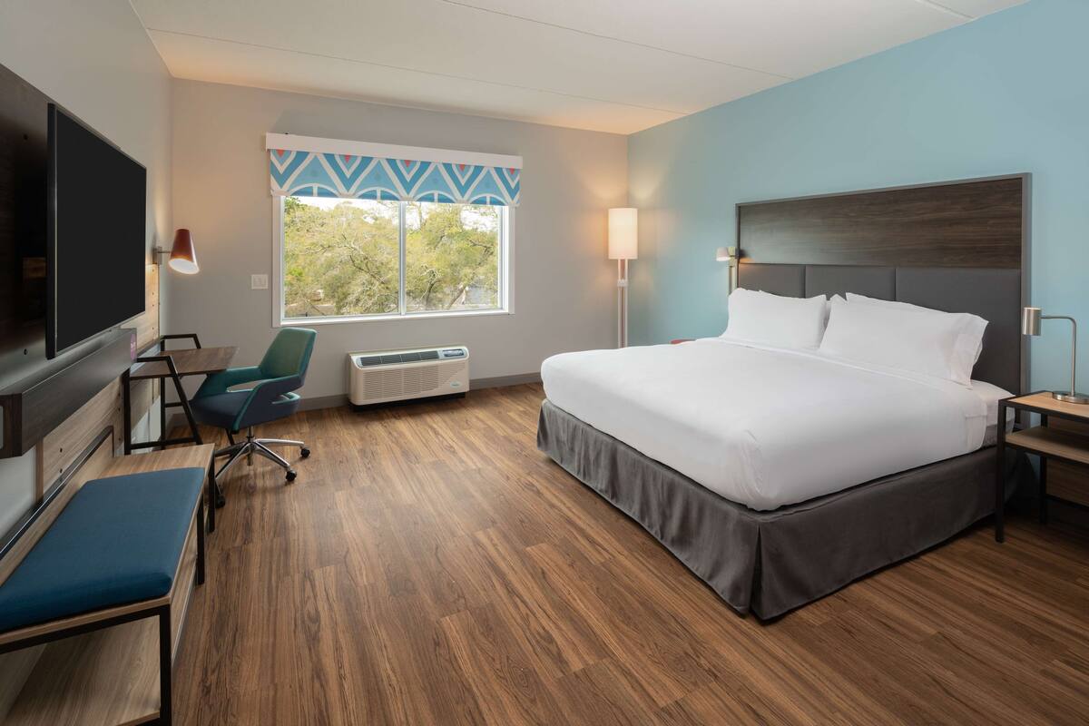 A hotel room with a large bed, a desk with a chair, a TV, and a window with a view of trees.