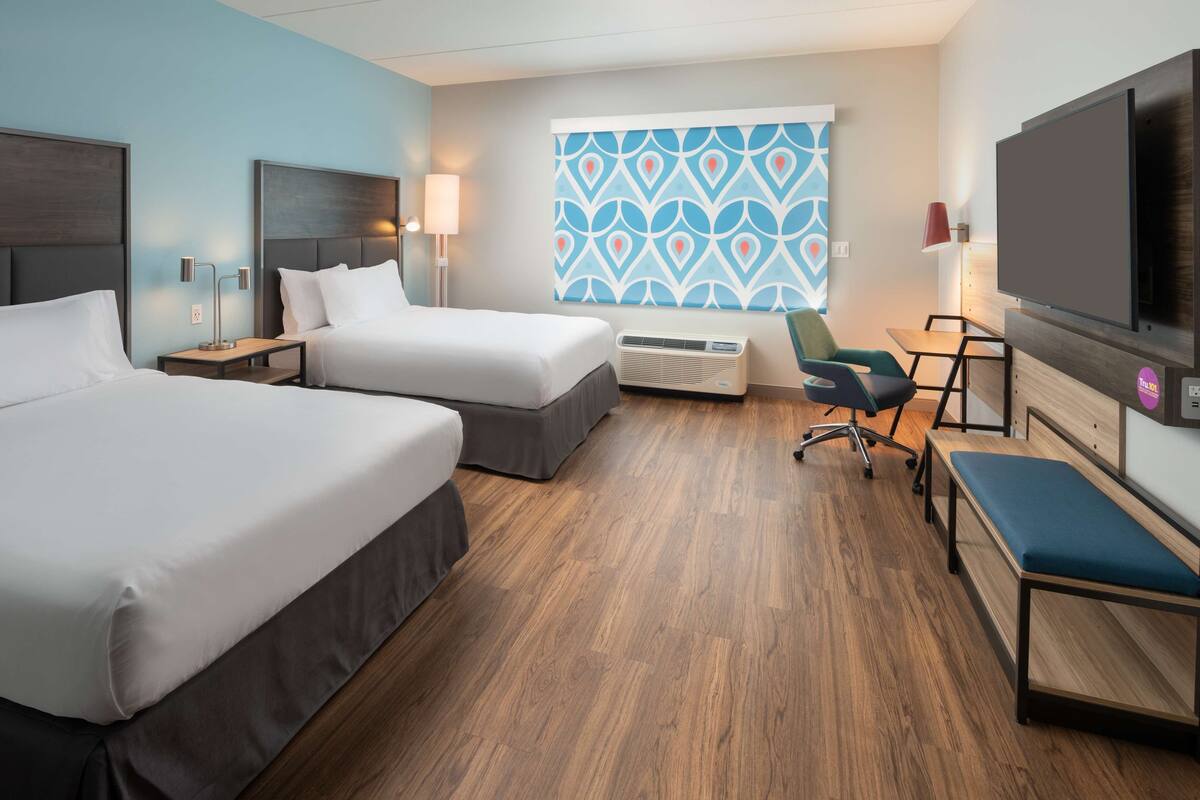 A hotel room with two beds, a desk, a chair, a TV, and a window with a geometric pattern.