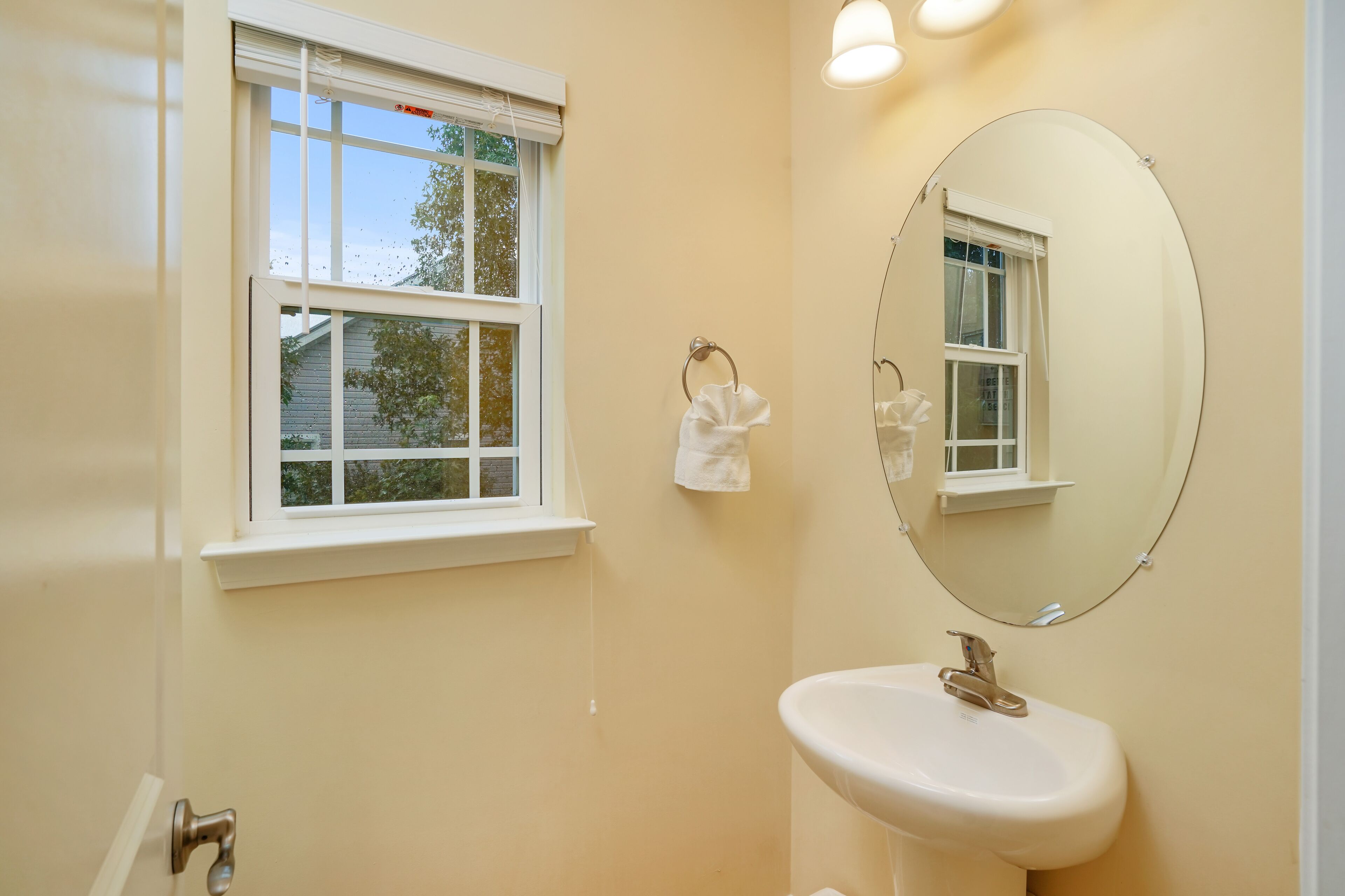 Un baño con un lavamanos blanco, un espejo grande y una ventana con vista a una casa y árboles.