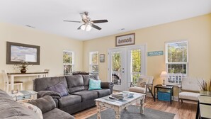 Ein Wohnzimmer mit grauem Sofa, einem Couchtisch und einem Deckenventilator. Zwei Türen führen nach draußen, und ein Schild an der Wand zeigt das Wort 'BEACH'.