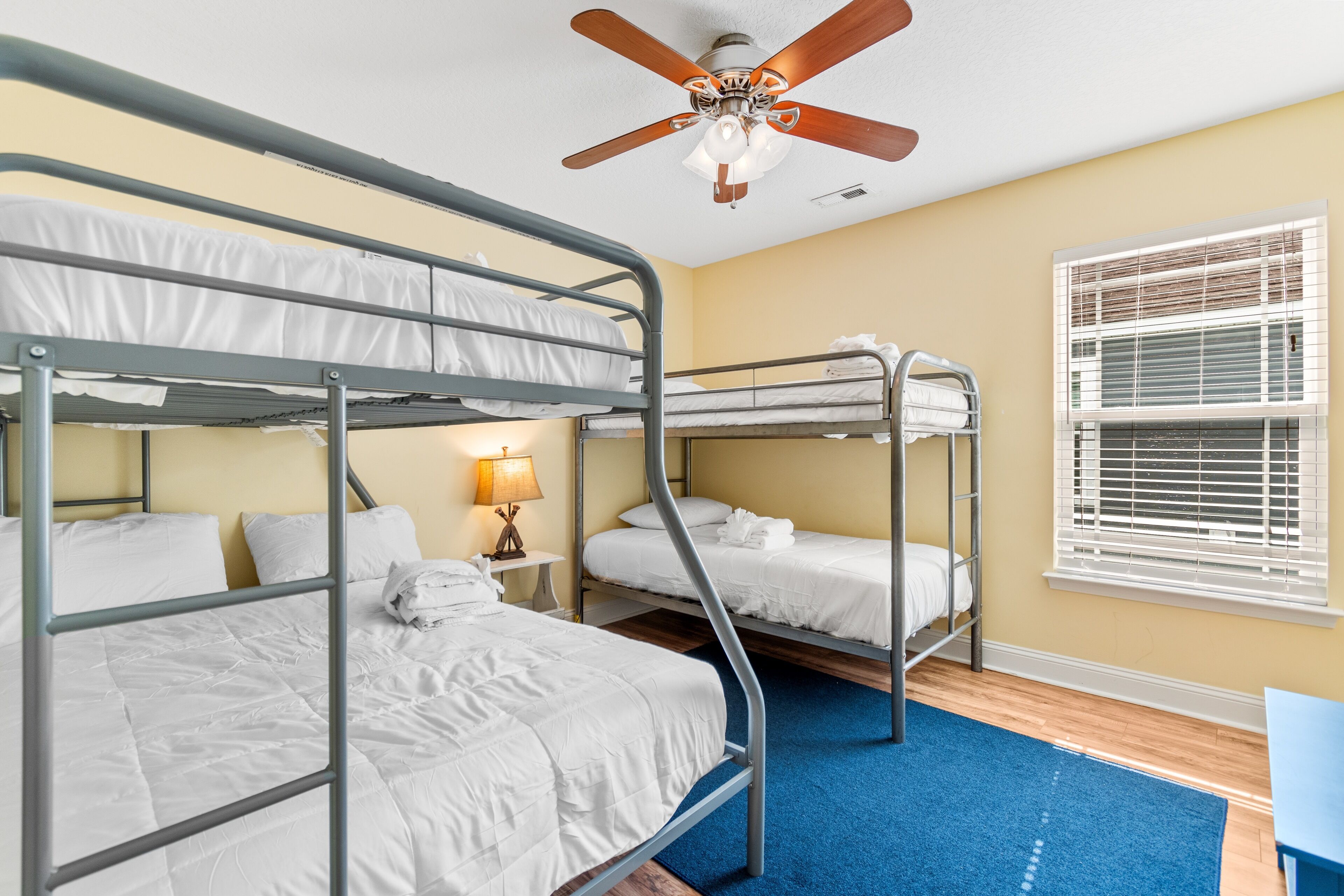 Une chambre avec des lits superposés, un ventilateur de plafond, une fenêtre avec des stores et un tapis bleu.