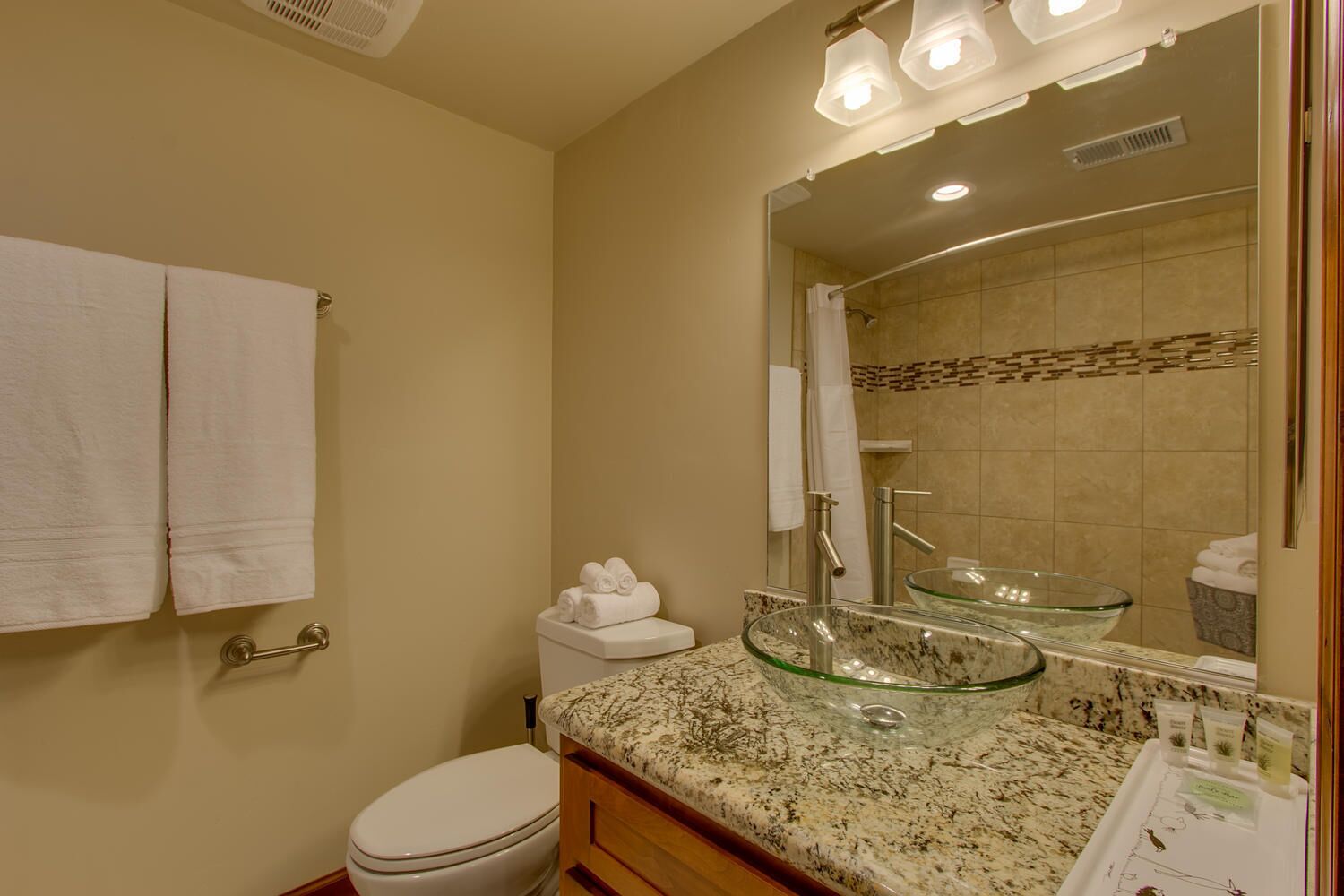 Une salle de bain comprenant un lavabo en verre, des toilettes, un porte-serviettes et un miroir avec éclairage intégré.