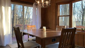 Una mesa de comedor de madera con sillas, una araña y ventanas amplias con vistas a árboles.