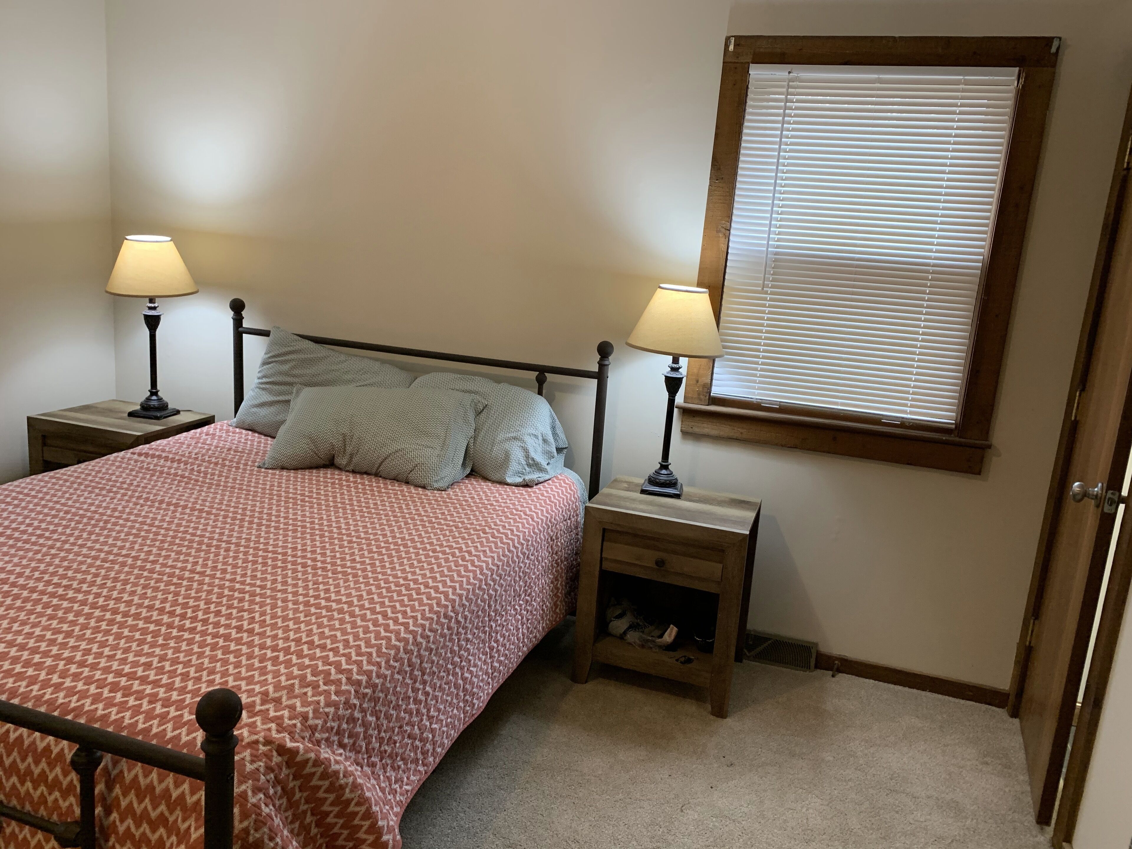 A bedroom with a bed, two bedside tables with lamps, a window with blinds, and a door.