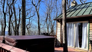 Una terraza de madera con una zona de barbacoa techada, una silla de madera, y una cabaña con techo verde y cortinas blancas.
