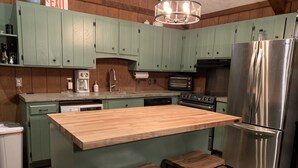 Una cocina con gabinetes verdes, un refrigerador de acero inoxidable, una isla de madera con taburetes, un horno empotrado y una cafetera.