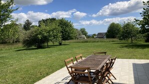 Patio esterno con tavolo e sedie in legno, circondato da prato e alberi.