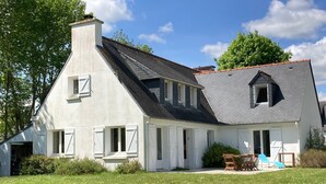 Una casa a due piani con tetto spiovente, pareti esterne bianche e un camino. C'è un piccolo portico con un tavolo e delle sedie, e una sedia blu all'esterno.