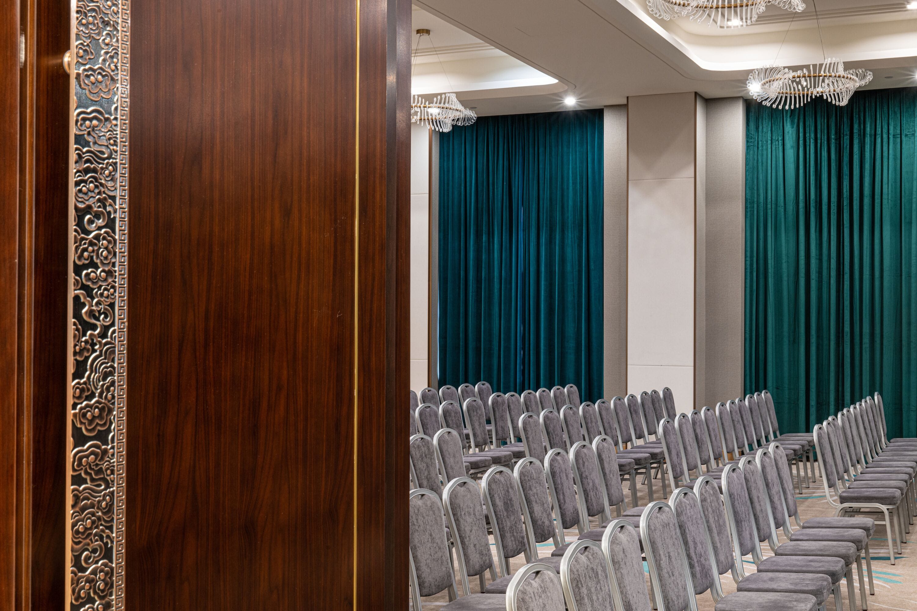 A conference room with rows of grey chairs, teal curtains, and a wooden door with intricate carvings.