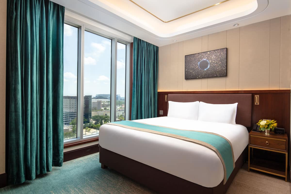 A hotel room with a large bed, a nightstand, a wall-mounted artwork, and a view of the city through large windows.
