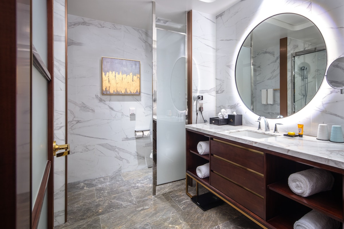 A modern bathroom with marble walls, a large mirror, a glass shower enclosure, and a vanity with towels.