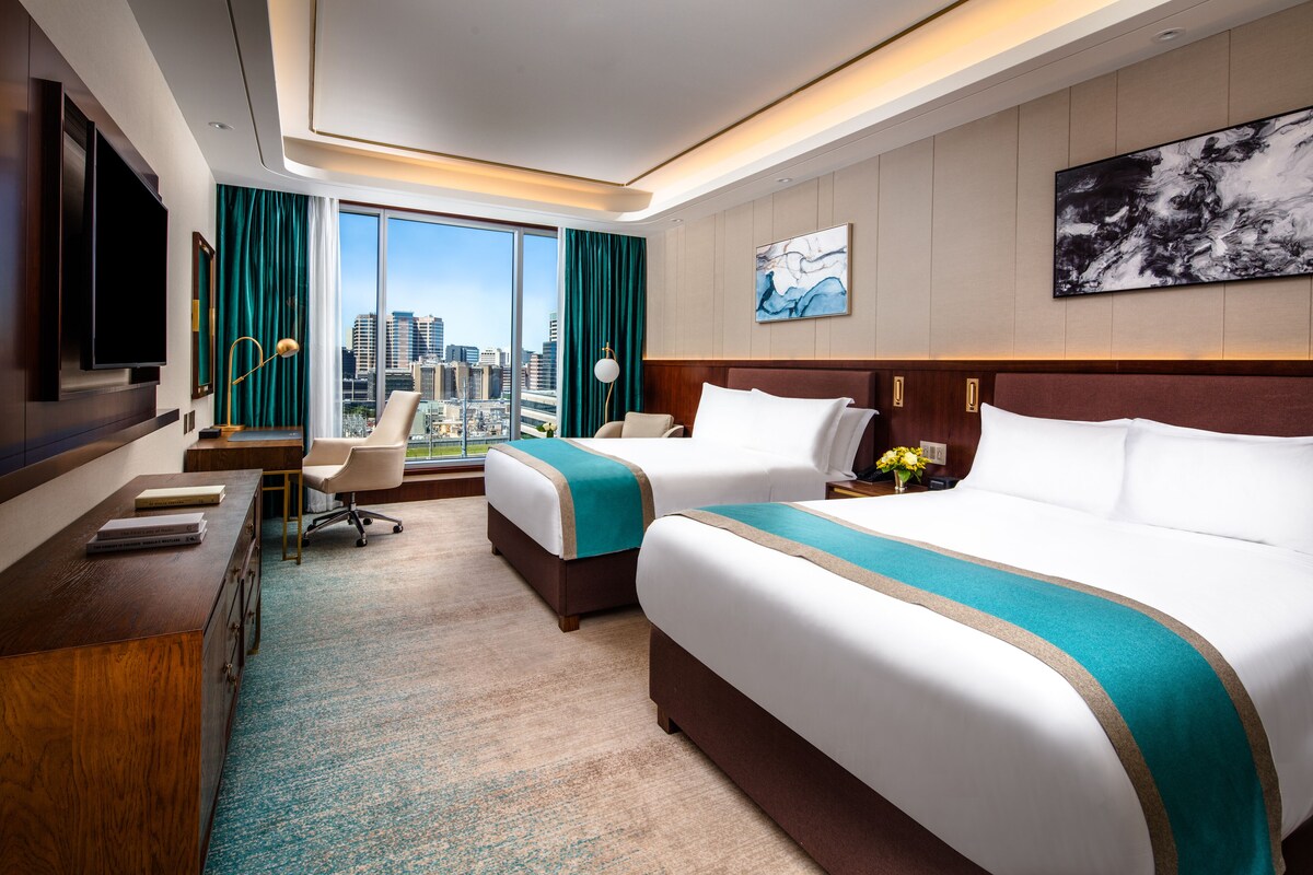 A hotel room with two beds, a desk, a chair, a TV, and a large window offering a city view.