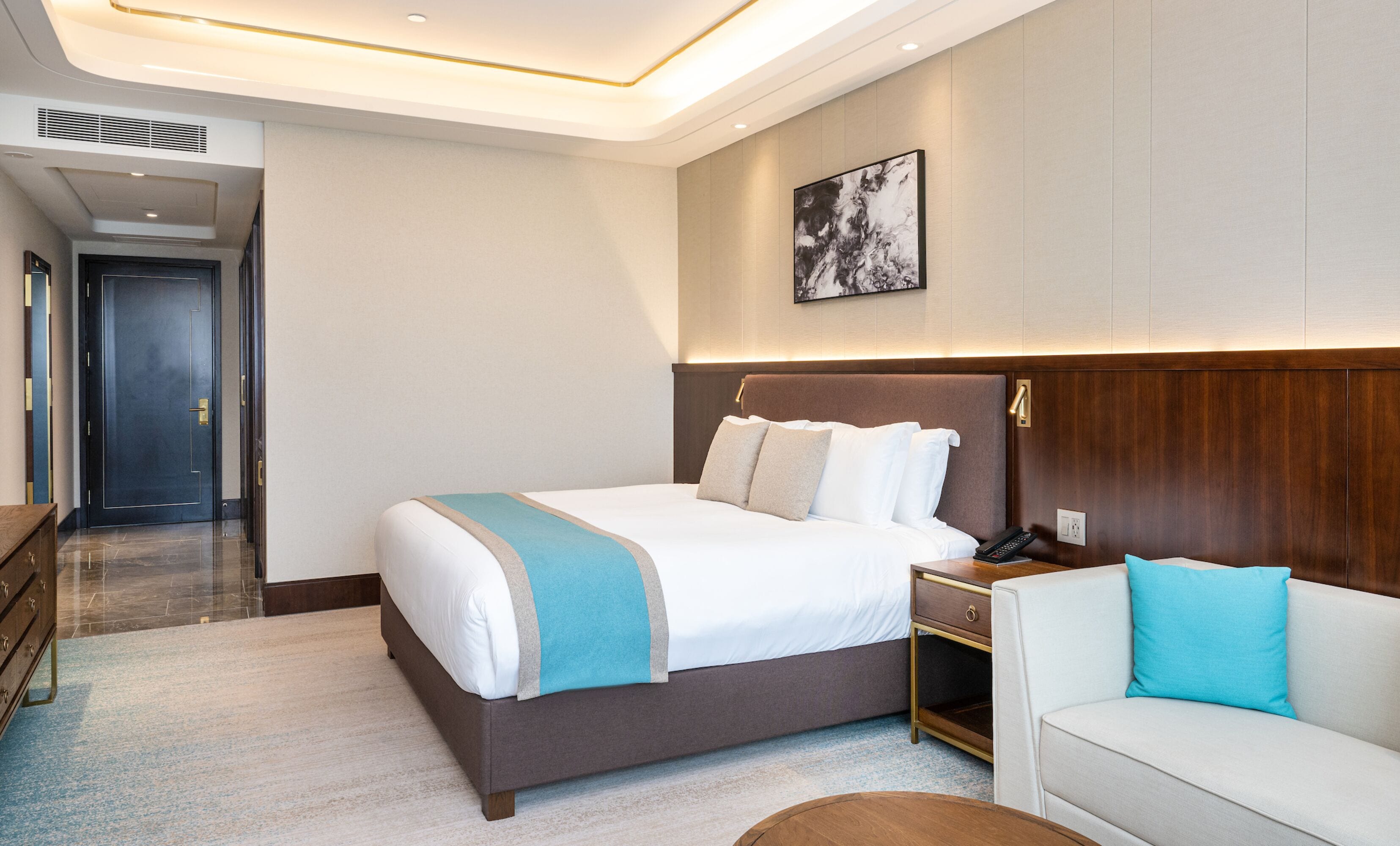 A modern hotel room with a large bed, a bedside table, a nightstand, a sofa, and a painting on the wall.