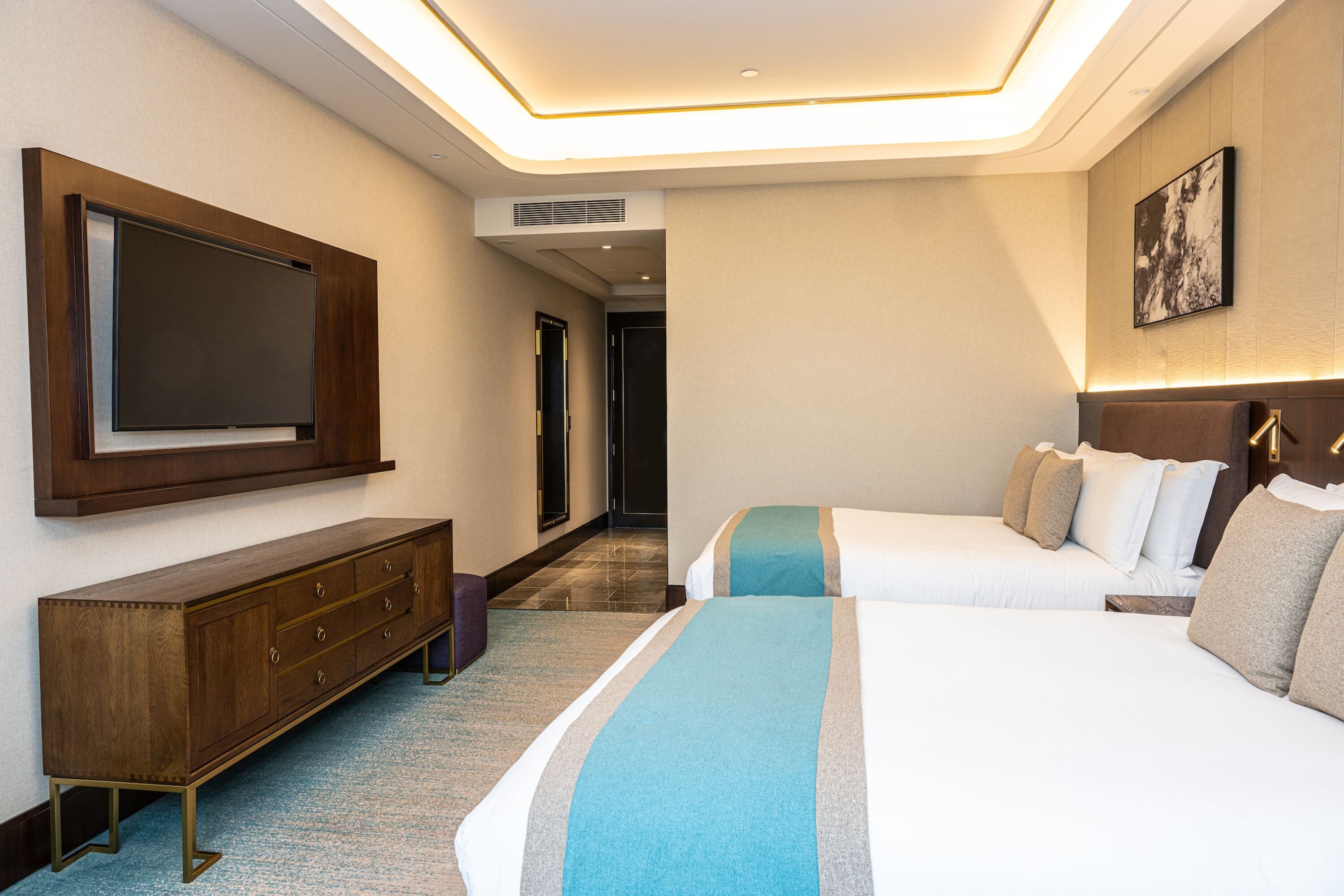 A hotel room with a large flat-screen TV mounted on the wall, a wooden console table, two beds with white and blue bedding, and a framed artwork on the wall.