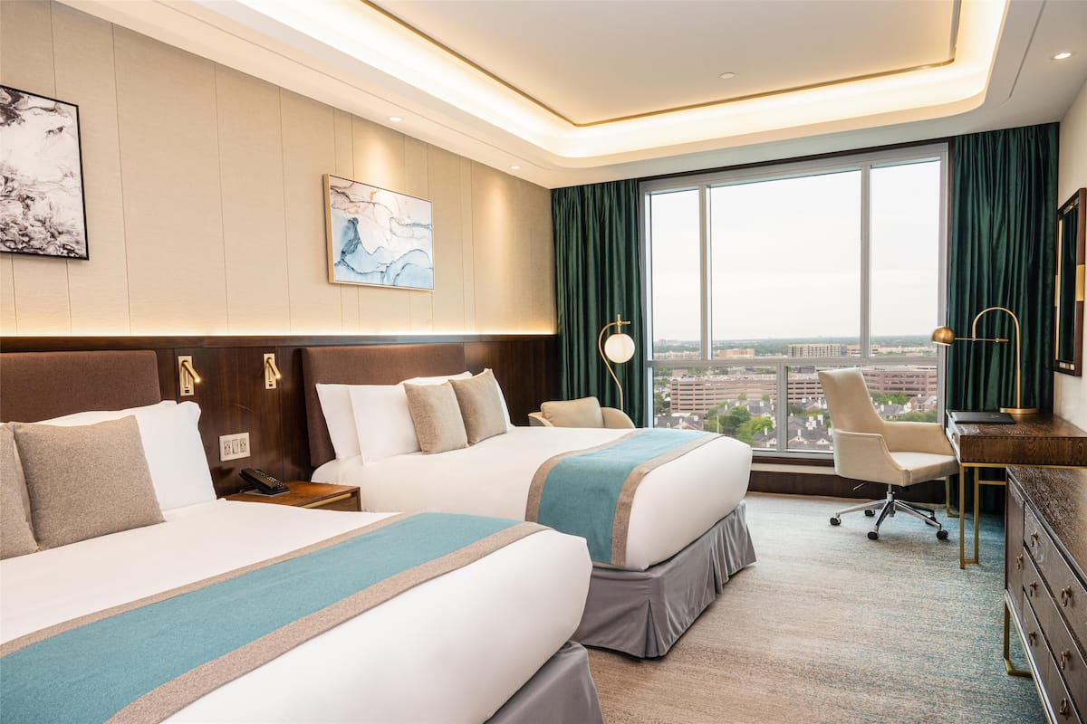 A hotel room with two beds, a desk, a chair, a large window with a view, and artwork on the walls.