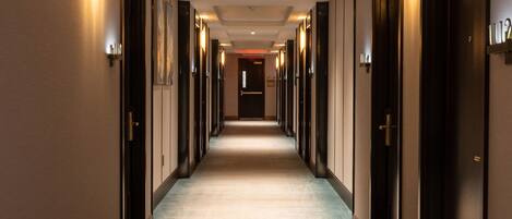A hotel corridor with doors on both sides, a carpeted floor, and wall-mounted lighting.