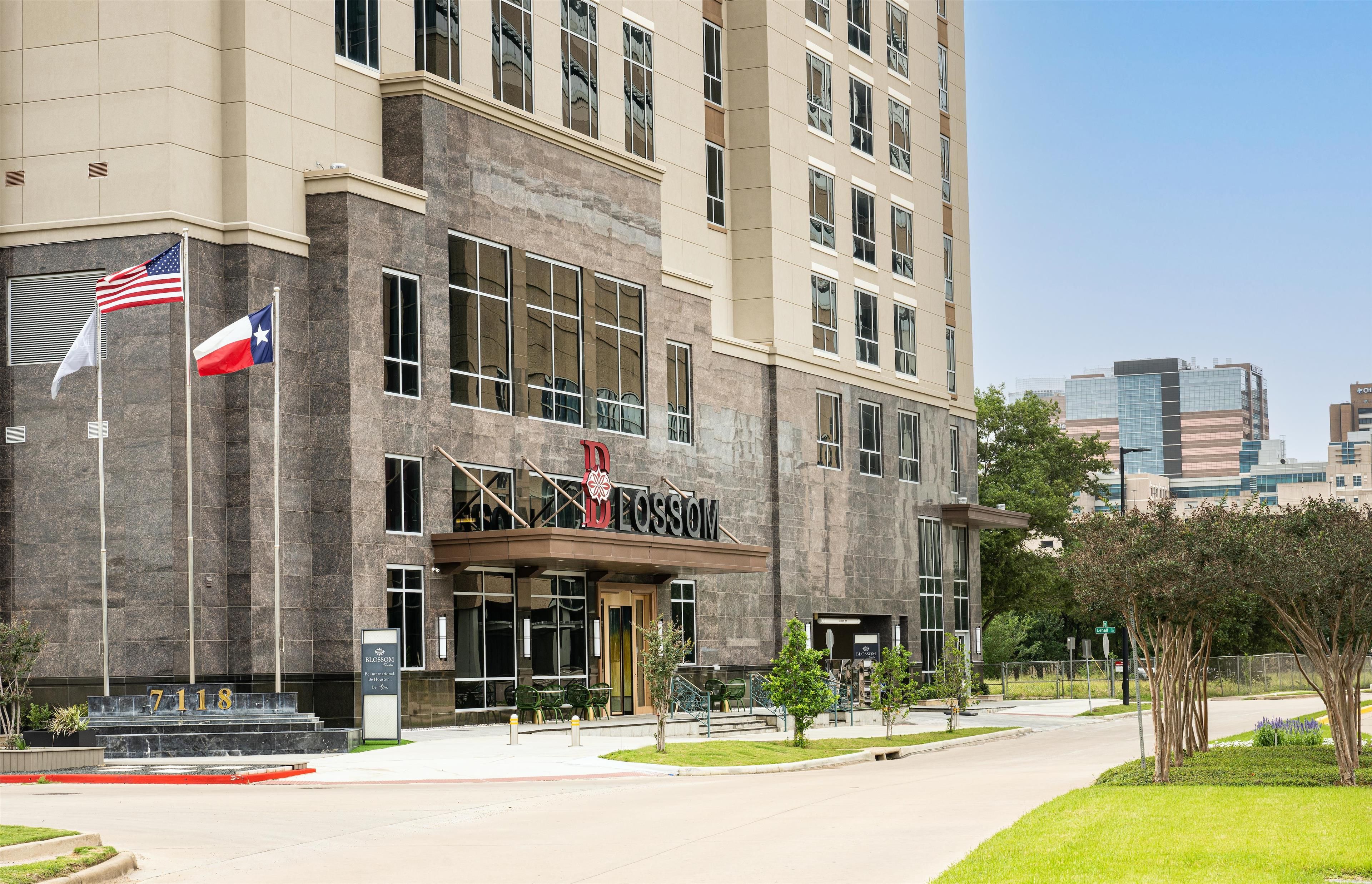 Um hotel com fachada de pedra, um mastro com a bandeira americana e uma placa com o nome 'Blossom Hotel'.