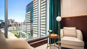 A modern hotel room with a curved glass wall, a view of a cityscape, a round side table with a plate of fruit, and two beige armchairs.