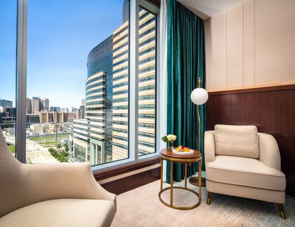 A modern hotel room with a curved glass wall, a view of a cityscape, a round side table with a plate of fruit, and two beige armchairs. - Blossom Hotel Houston, Curio Collection by Hilton (Houston)