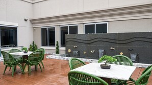 An outdoor patio area with green wicker chairs and a marble table, surrounded by a modern building with large windows and decorative wall panels.