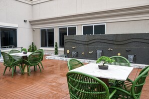 An outdoor patio area with green wicker chairs and a marble table, surrounded by a modern building with large windows and decorative wall panels. - Blossom Hotel Houston, Curio Collection by Hilton (Houston)