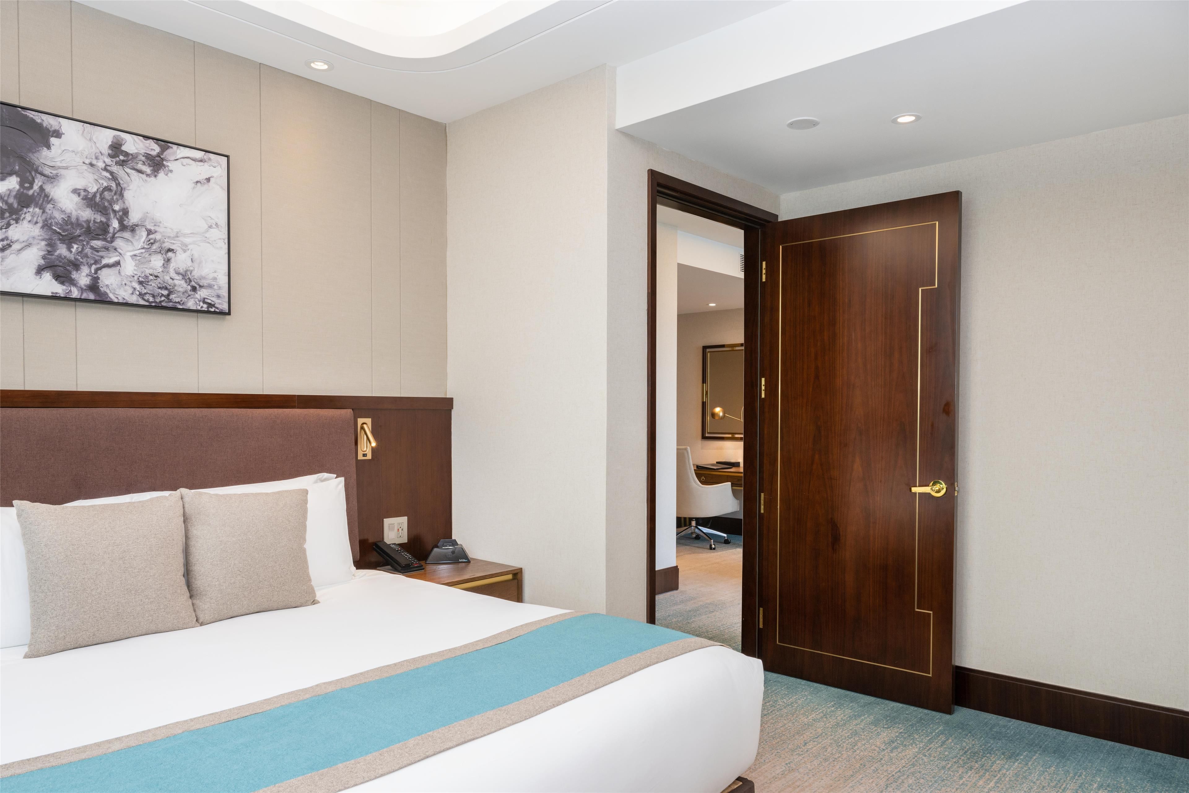 A hotel room with a large bed, a bedside table with a phone, a wall-mounted artwork, and a wooden door.