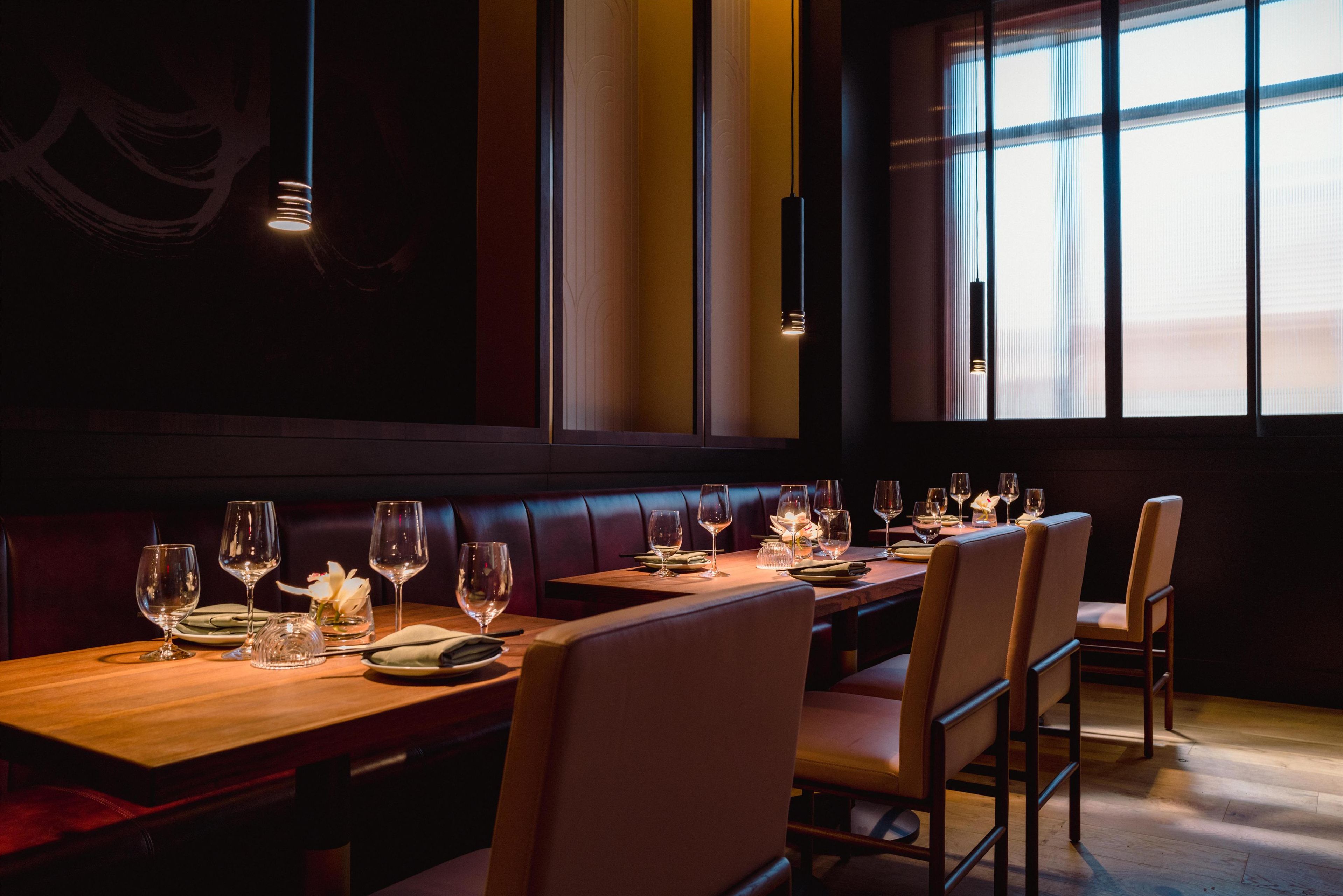 A restaurant with a long wooden table set for a meal, leather booths, and large windows allowing natural light.