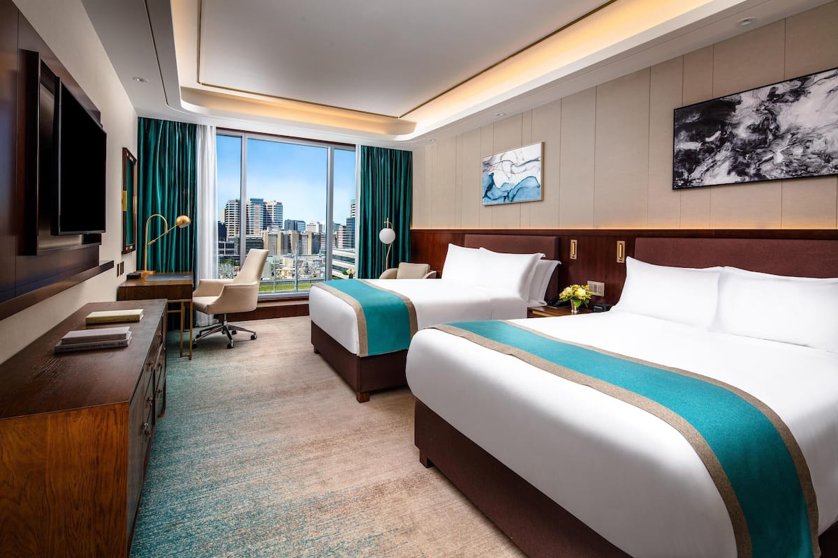 A hotel room with two beds, a desk, a TV, and a large window with a city view.