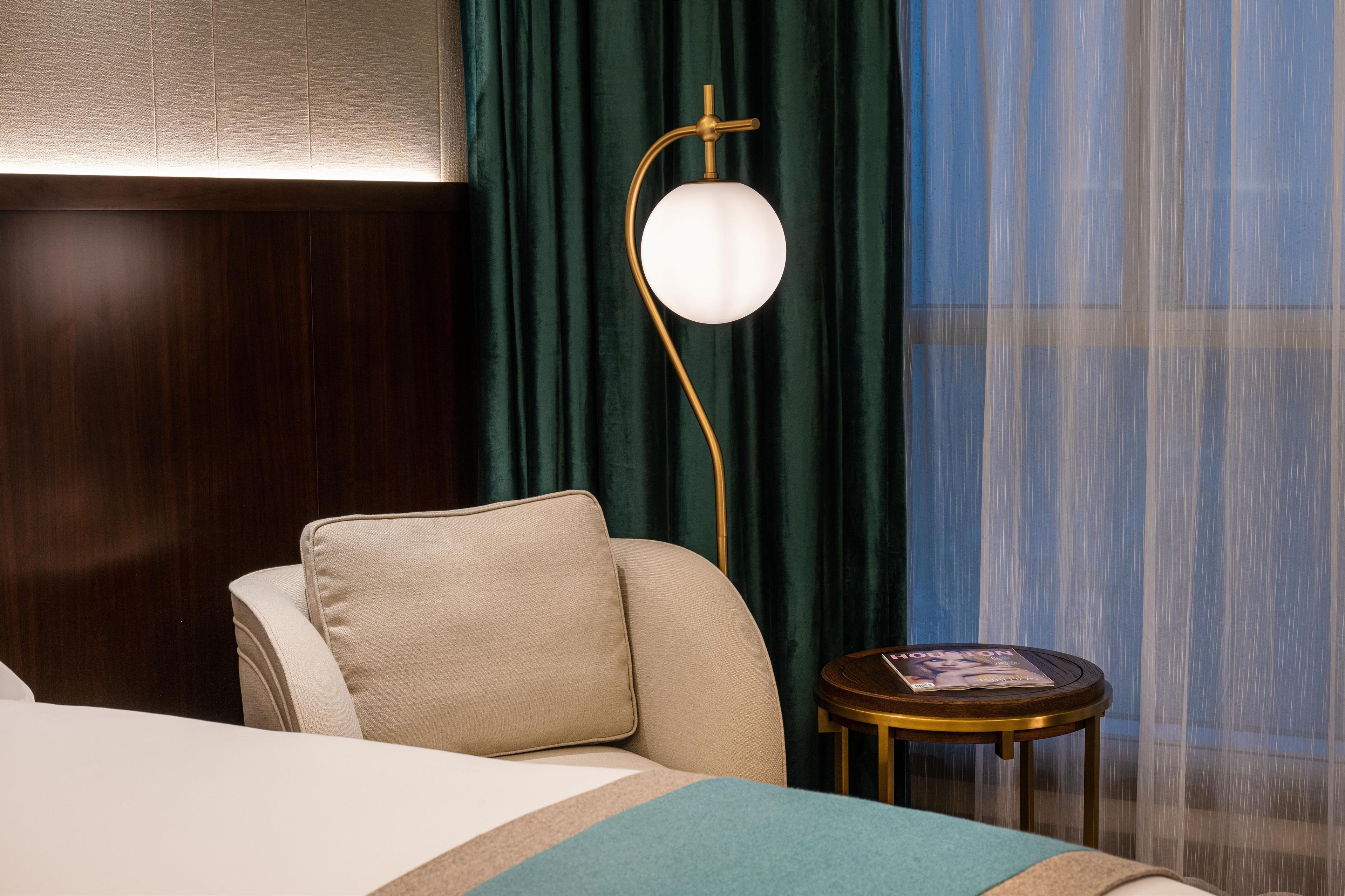 A hotel room with a bed, a round side table with a magazine, a floor lamp, and a view of the outside through sheer curtains.